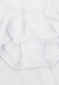 Ein weißer, transparenter Stoff liegt über einem glatten, weißen Kleidungsstück und zeigt eine weiche Textur und nahtlose Kanten, mit geschwungenen Ausschnitten für ein figurbetontes Design.