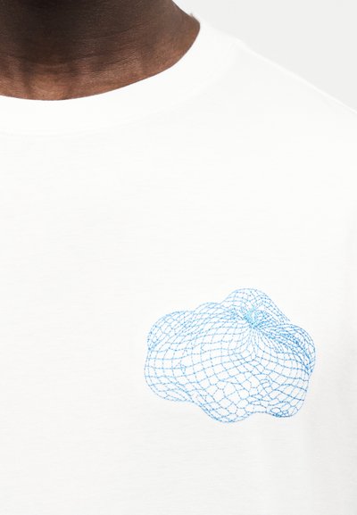 Κοντινό πλάνο ενός ατόμου που φοράει λευκό πουκάμισο με μικρή μπλε αφηρημένη κεντημένη απεικόνιση σε σχήμα δικτύου στην περιοχή του στήθους.