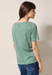 Haut à manches courtes, vert clair, avec un motif en relief de labyrinthe et un petit détail en forme de trou de serrure au dos. Conçu pour un ajustement décontracté.