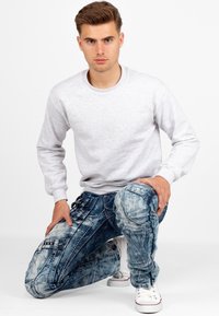 Sweatshirt grigio a girocollo abbinato a jeans blu strappati con tasche e dettagli di cucitura. Il modello è in ginocchio con una postura casual.