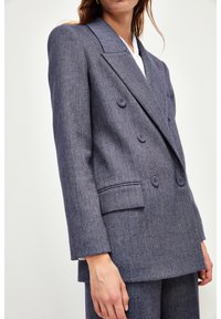 Blazer de doble botonadura en azul marino con un patrón de pata de gallo, que destaca por sus amplios solapas, hombros estructurados y bolsillos frontales con solapa.
