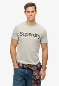Lys grå bomulds T-shirt med sort "Superdry" logo på tværs af brystet. Sammen med blå jeans og en rød ternet skjorte bundet omkring livet.