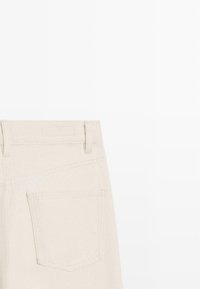 Un pantalon en coton beige clair présente une taille haute, une coupe droite et une seule poche arrière avec des coutures discrètes le long des coutures.