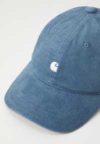 Boné de corduroy azul com superfície texturizada, aba curva e etiqueta de logo branca. Apresenta costuras cozidas e ilhós de ventilação.