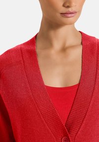 Rode cardigan met ribbelstructuur en een diepe V-hals. Voorzien van een knoopsluiting en gelaagd over een rode tanktop.