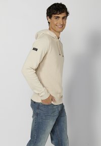 Pull à capuche beige avec un motif texturé et un cordon de serrage. Possède une patch sur la manche gauche. Associé à un jean bleu.
