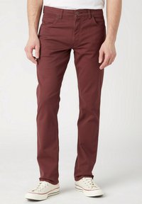 Pantalones de algodón burdeos con un corte ajustado, diseño de cinco bolsillos y cierre de botón, combinados con zapatillas blancas. Textura suave y estilo casual.