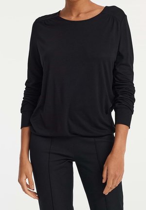 Kobieta ubrana w gładką czarną bluzkę z długim rękawem i dopasowane czarne spodnie, stojąca na tle jasnego tła.