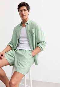 Camicia button-up in lino verde chiaro con tasca frontale, abbinata a pantaloni corti coordinati. Sotto, è visibile una canotta bianca. Design semplice.