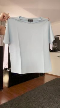 Tricou scurt cu mânecă scurtă, de culoare albastru deschis, ținut întins de două mâini în interior, peste un podea din lemn și un covor gri.