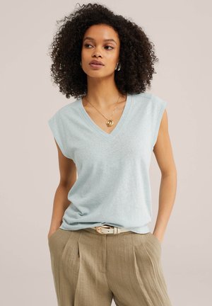 Göndör hajú nő világoskék ujjatlan V-nyakú felsőben, bézs nadrágban övvel és arany spirál medállal.