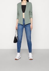 Skuren grön blazer med långa ärmar, öppen framsida, i kombination med en svart kort topp, blå jeans, vita sneakers och en svart handväska.