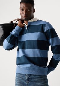 Polo à manches longues bleu clair et foncé à rayures avec un col blanc. Le modèle porte un sac noir et un jean en denim bleu.