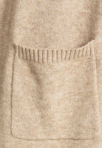 Béžový pletený svetr s měkkou texturou, s výraznou přední kapsou a žebrovanými detaily podél okraje kapsy.