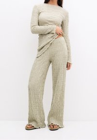 Vrouw in een beige, textuurgegeven, lange mouwen top en wijdvallende broek, die de zoom iets omhoog heft, met open teentoegankelijke beige sandalen op een witte achtergrond.