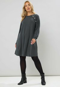 Abito grigio a maniche lunghe a coste con una vestibilità rilassata, caratterizzato da tre bottoni decorativi sulla spalla, abbinato a stivali neri alla caviglia.