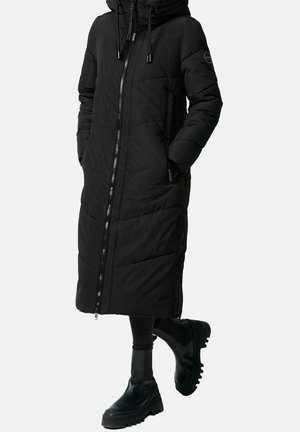 Long manteau noir matelassé avec fermeture éclair à l'avant et capuche, porté avec des leggings noirs et des bottes à plateforme épaisses noires. Mains dans les poches du manteau.
