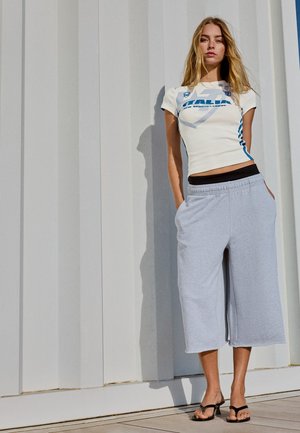 Mladi žena s dugom plavom kosom u uskoj bijeloj sportskoj majici "Italia", svijetlosivim širokim skraćenim hlačama i crnim sandalama na petu.