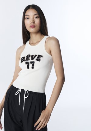 Jonge vrouw met lang donker haar, die een wit mouwloos shirt draagt met de tekst "REVE 77" en losse zwarte broek met witte trekkoorden.