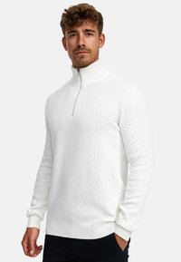 Ung mann med lys brunt krøllete hår som har på seg en hvit teksturert genser med kvart-glidelås og svarte bukser mot en ensfarget bakgrunn.