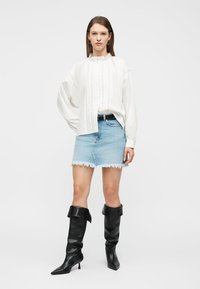 Γυναίκα που φοράει λευκό μπλουζάκι με μακριά μανίκια, ανοιχτό μπλε σκισμένη τζιν φούστα, μαύρη ζώνη και μαύρες μπότες με τακούνι μέχρι το γόνατο, στεκόμενη σε λευκό φόντο.