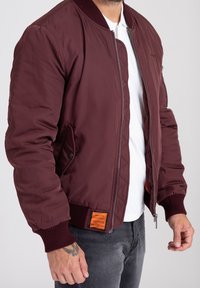 Bordeaux bomberjacka med ribbat krage och muddar, dragkedjestängning och sidofickor. Har en orange etikett på nedre vänstra sidan.