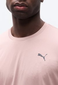 Κοντινή εικόνα ενός ατόμου που φοράει ένα ανοιχτό ροζ πουκάμισο με ένα μικρό, σκοτεινό λογότυπο Puma στην αριστερή πλευρά του στήθους.