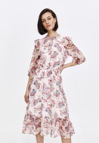 Model pozuje pewnie w pastelowej sukience z wzorami paisley i motyli, z falbaniastymi rękawami i zwiewnym dołem.