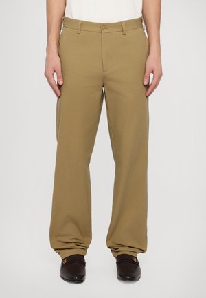 Khaki puuvillahousut rentolla leikkauksella, varustettuna nappisuljennuksella ja vyölenkeillä. Yhdistetty tummiin loafer-kenkiin ja valkoiseen paitaan.