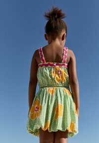 Robe à carreaux verts avec un motif floral jaune, bretelles festonnées roses et taille cintrée, vue de dos contre un ciel bleu clair.