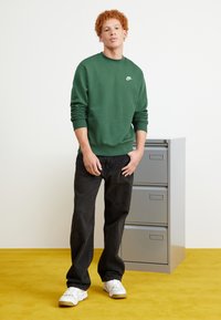 Sudadera verde con puños y dobladillo acanalados, que presenta un pequeño logotipo blanco de Nike. Combinada con unos vaqueros negros holgados y zapatillas blancas sobre una alfombra amarilla.