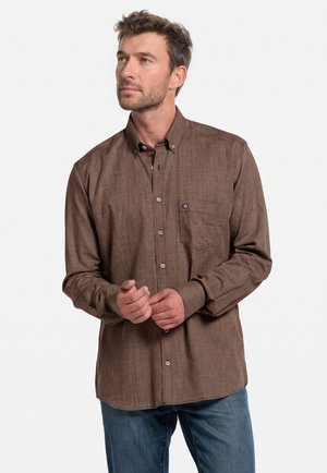 Mann trägt ein braunes Hemd mit Knöpfen und Brusttasche und blaue Jeans, blickt leicht nach links mit verschränkten Händen.