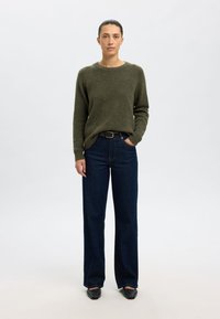 Olivgrüner Wollpullover und dunkelblaue Jeans mit weitem Bein, kombiniert mit schwarzen Slippern. Einfaches Design ohne sichtbare Muster oder Verzierungen.