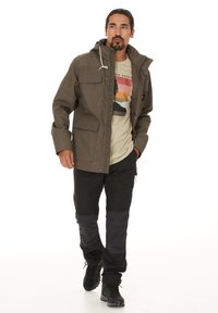 Man som bär en brun huva jacka över en beige t-shirt med grafik, svarta utomhusbyxor och svarta vandringskängor, går med ena handen i fickan.