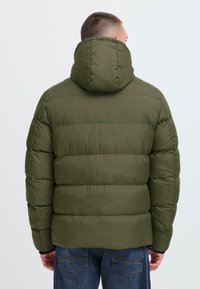 Man wearing olive green padded hooded jacket and blue jeans, shown from the back against a plain light background.