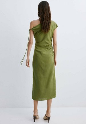 Žena s dugom valovitom kosom u maslinasto zelenoj midi haljini na jedno rame i bež štiklama, stoji na jednostavnoj svijetloj pozadini.