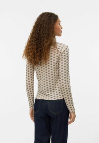 Top de manga larga ajustado en color beige con un patrón floral negro, fabricado en un tejido suave. Combinado con jeans azul oscuro, que muestran un dobladillo curvado en la parte posterior.