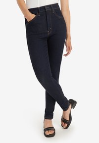 Sötét mosású farmernadrág magas derékkal, slim fit szabással és finom narancssárga varrással. A modell fekete, pántos magassarkú szandállal párosítja őket.
