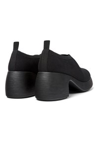 Chaussures noires avec une tige en tissu texturé et un talon épais incurvé, dotées de languettes à l'arrière pour un enfilage facile.