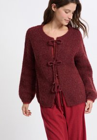 Cardigan in magenta lavorato a maglia con scollo rotondo, tessuto testurizzato, chiusure legate e maniche a palloncino. Abbinato a pantaloni a scacchi rossi.
