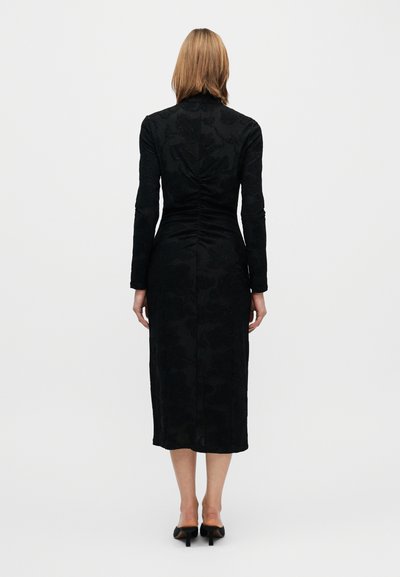 Fekete, hosszú ujjú ruha, testhez simuló sziluettel, texturált virágmintával, és középhosszúságú, borjúig érő szabással. Hátsó hasítékkal és finom ráncolással díszített.