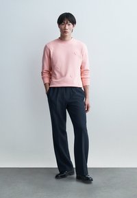 Mladi moški stoji z rokami v žepih, oblečen v roza jopico z vzorci src, črne hlače in črne čevlje, na preprostem ozadju.