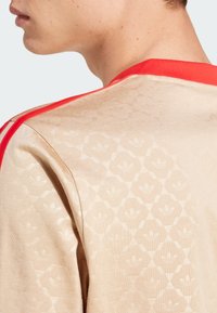 Beige sporta krekls ar teksturētu rakstu, sarkanu apkakli un plecu svītrām. Materiāls izskatās mīksts un gluds, izceļot zīmola detalizāciju.