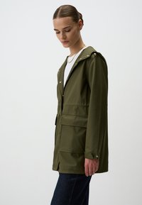 Olive zielona wodoodporna kurtka z kapturem, z guzikami z przodu, dwoma przednimi kieszeniami i gładką fakturą. Model w niej stoi z boku.