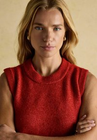 Roter ärmelloser Pullover mit rundem Ausschnitt und strukturiertem Strick. Glattes Material zeigt einen leichten Schimmer. Modell mit verschränkten Armen vor neutralem Hintergrund.