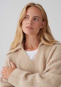 Maglione beige a maglia con scollo a V e vestibilità ampia. Presenta una texture a costine e spalle scese. Indossato sopra una camicia bianca a maniche lunghe.