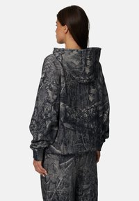 Huzarowa bluza w kamuflażu z ciemnoszarym i czarnym wzorem leśnym, luźny krój oraz kieszenią z przodu i kapturem ze sznurkiem.