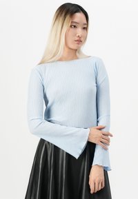 Mladá žena s dlouhými blond vlasy, oblečená v světle modrém dlouhém rukávu s texturou a černé skládací sukni, na bílém pozadí.