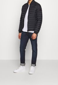 Man i svart vadderad jacka över vit t-shirt, mörka uppvikta jeans, vita strumpor och vita sneakers mot en plain bakgrund.