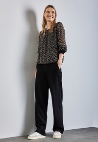Schwarze, transparente Bluse mit langen Ärmeln und geometrischem Muster, kombiniert mit schwarzen, weiten Hosen und cremefarbenen Sneakers. Glatte Texturen und lässige Passform.